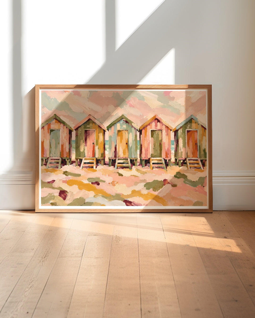 A framed art print depicting a row of colorful beach huts in a soft, impressionistic style, leaning against a white wall on a light wood floor with natural window shadows.