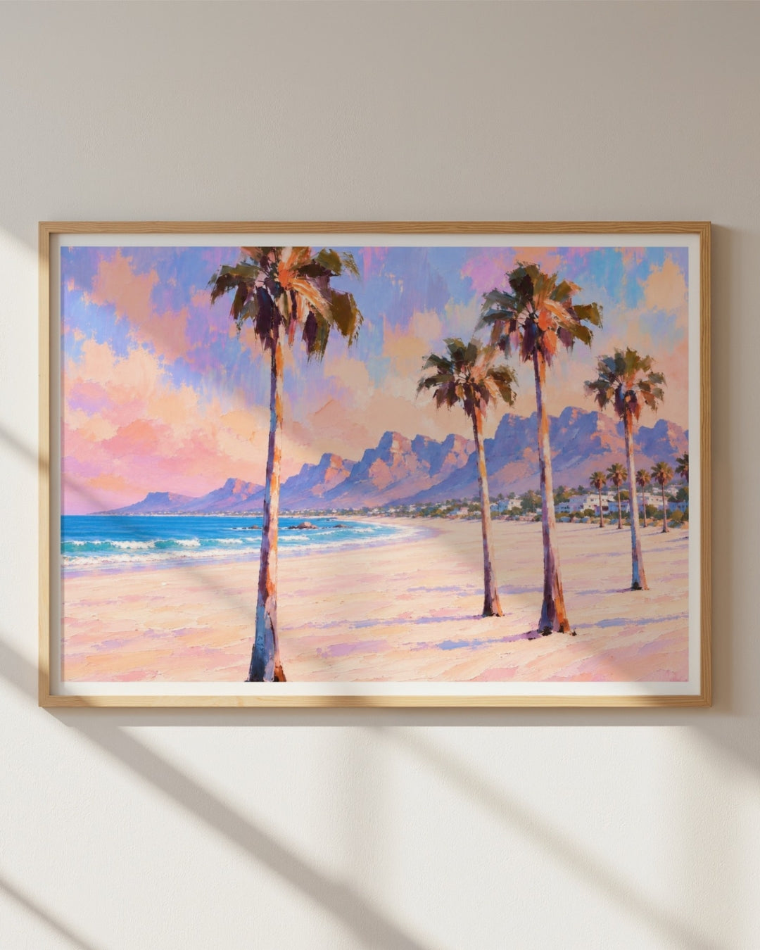 A framed beach landscape painting featuring tall palm trees, blue ocean water, distant mountains, and a pastel sky, displayed on a light wall with soft window shadows.