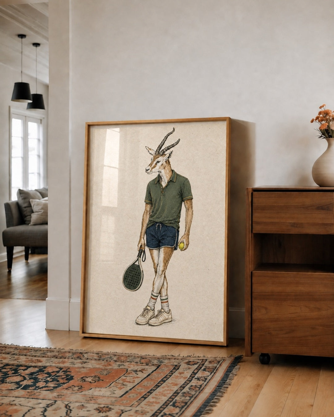 A framed art print featuring a whimsical illustration of a male springbok antelope dressed in a green polo shirt, blue shorts, and holding a tennis racket and tennis ball, leaning against a neutral wall in a modern living room setting.