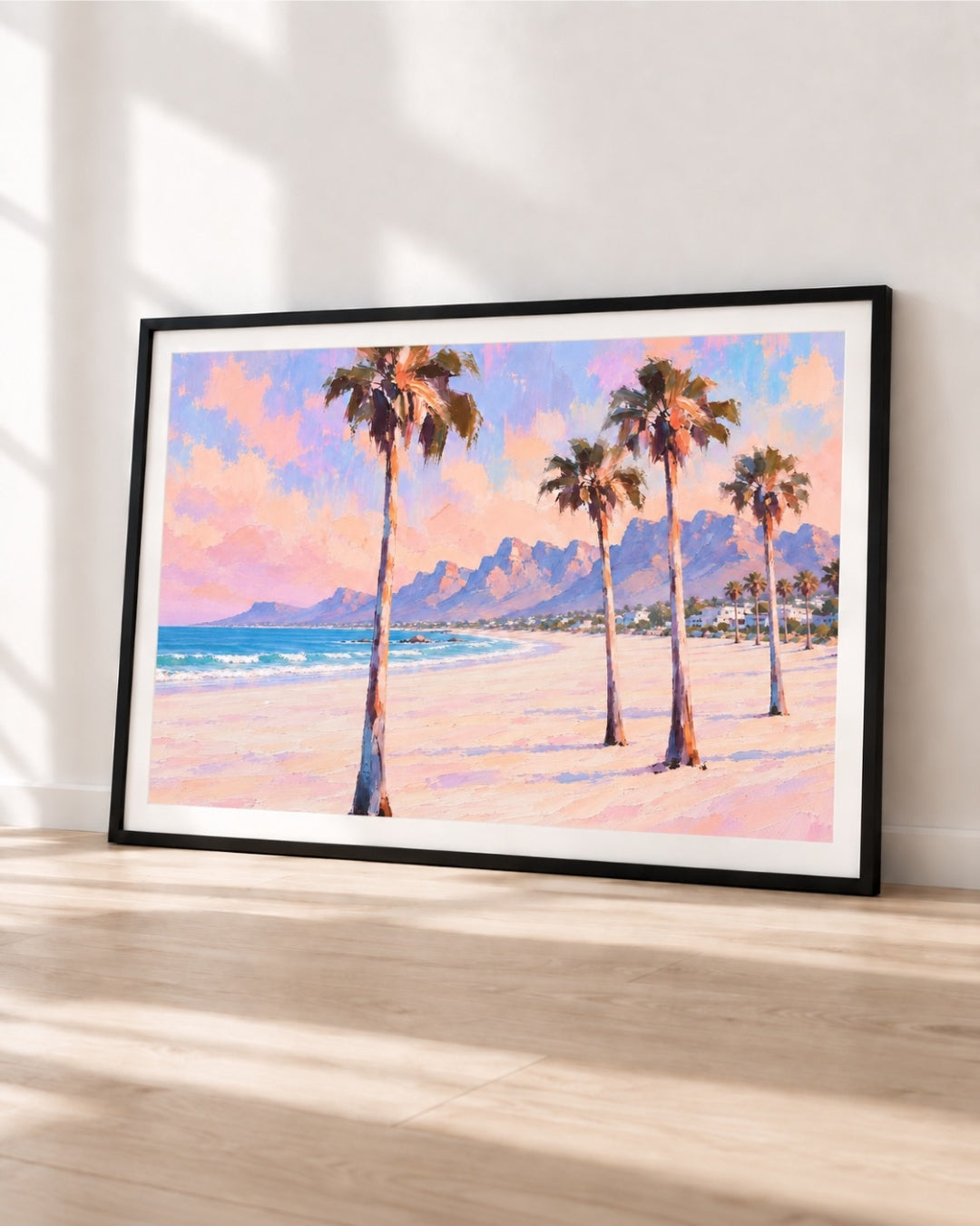 A framed beach landscape print leaning against a white wall on a light wood floor. The artwork shows tall palm trees on a sandy shore with blue ocean water and mountains in the background under a pink and blue sky.