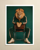 A framed art print featuring a stylized portrait of a lion with a human face, seated on a wooden chair and wearing a dark green tracksuit with beige stripes, against a dark teal background. The print is displayed on a light beige wall.
