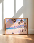 A framed landscape painting of a tropical beach with palm trees, ocean, and mountains, leaning against a white wall on a light wood floor with dramatic window shadows.