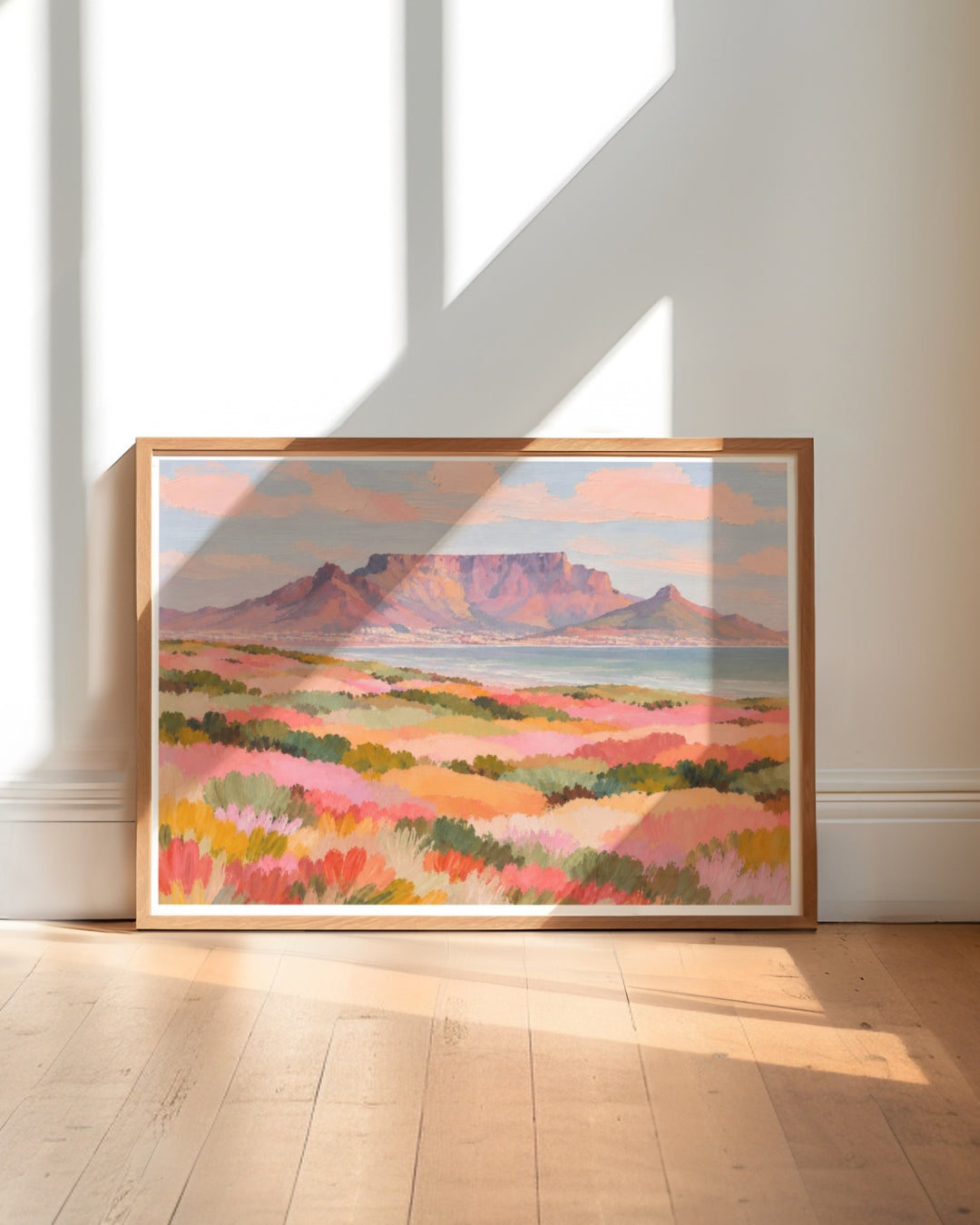 A framed landscape art print depicting a mountain range and colorful fields, leaning against a white wall on a light wood floor with natural window shadows.