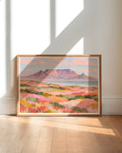 A framed landscape art print depicting a mountain range and colorful fields, leaning against a white wall on a light wood floor with natural window shadows.