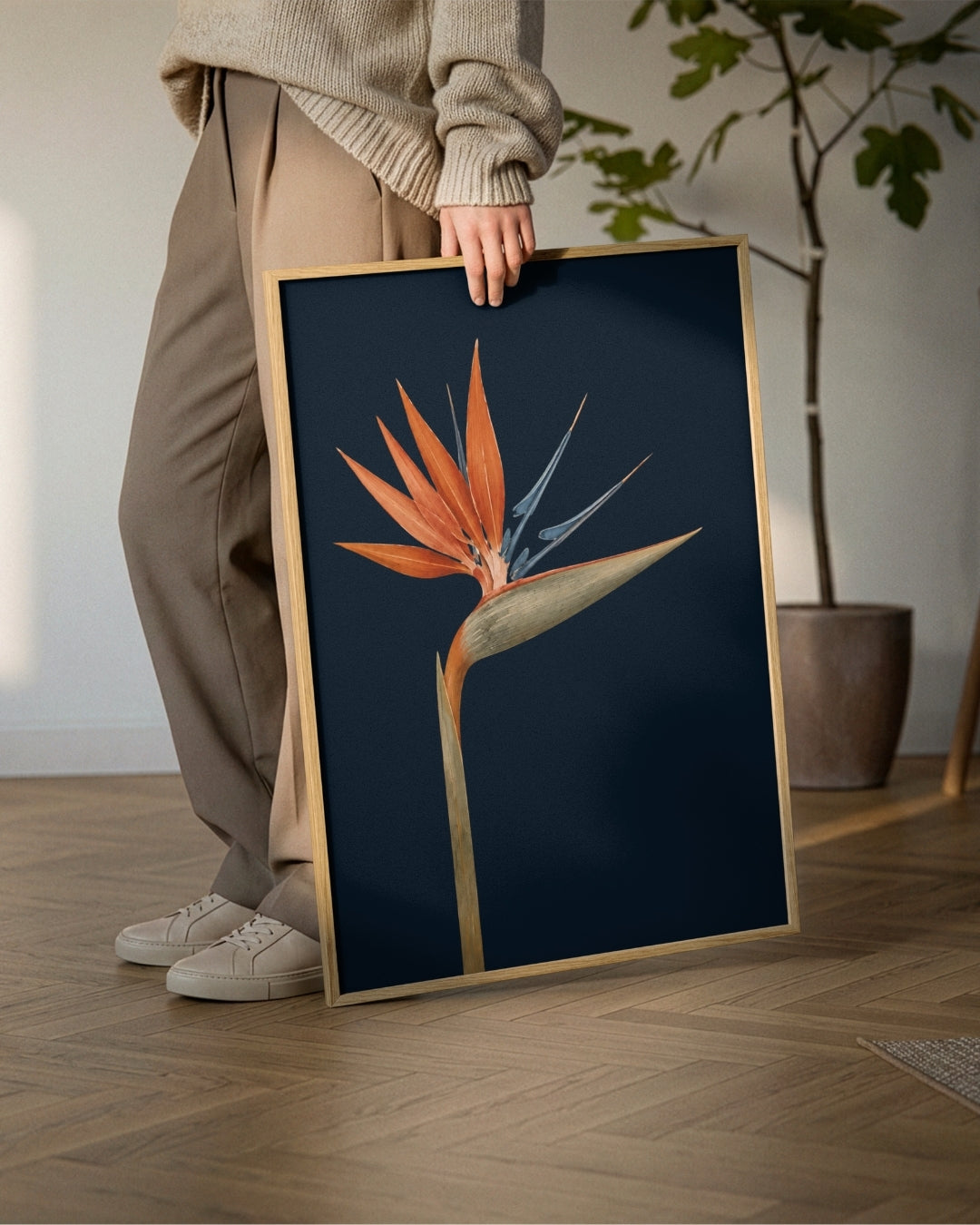 A person in a beige sweater and brown pants holds a framed botanical art print featuring an orange and blue bird of paradise flower on a dark navy background, set in a modern room with a wooden floor and a potted plant.