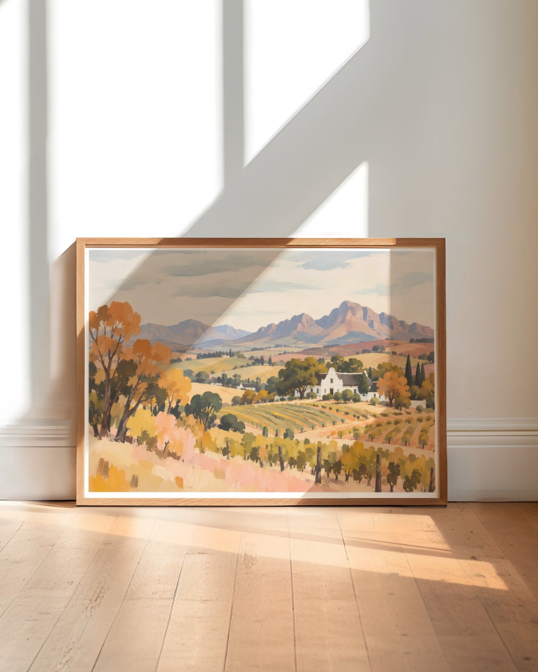 A framed landscape art print leaning against a white wall on a light wood floor, showing a vineyard scene with autumn trees, a white farmhouse, and mountains in the background.