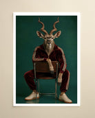 A framed art print featuring a surreal portrait of a kudu antelope dressed in a dark red velour tracksuit with white stripes, seated on a chair against a dark teal background. The print is displayed on a light beige wall.