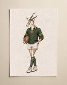 A vertical art print featuring a whimsical illustration of a springbok antelope dressed in a vintage rugby jersey, shorts, and socks, holding a rugby ball. The print is displayed on a neutral beige wall.