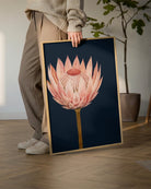 A person in a beige sweater and brown pants holds a framed art print featuring a pink protea flower on a dark navy background. The print is framed in light wood and shown in a modern room setting with a wooden floor and a potted plant in the background.