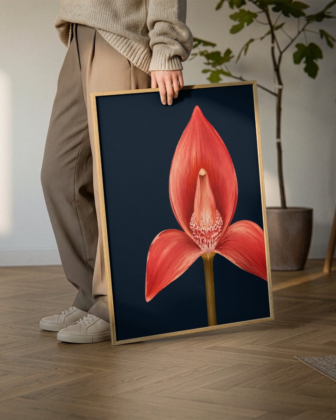 A person in beige sweatpants and white sneakers holds a framed botanical art print of a red-orange flower on a dark navy background, set in a modern room with a wooden floor and a potted plant.