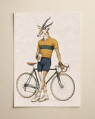 A vintage-style art print featuring a gazelle dressed as a cyclist with a road bike, shown hanging on a light beige wall.