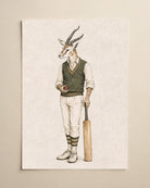A vertical art print featuring a whimsical illustration of a gazelle dressed in vintage cricket attire, holding a bat and wearing a cricket ball. The print is displayed on a neutral beige wall.