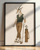 A framed art print featuring a whimsical illustration of a male springbok gazelle dressed in golf attire, standing next to a golf bag. The print is displayed in a black frame on a light-colored wall above a wooden floor, with soft diagonal shadows cast across the print.
