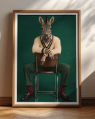 A framed art print featuring a zebra dressed in a cream sweater, green trousers, and red socks, seated on a chair against a dark green background. The print is displayed in a wooden frame leaning against a beige wall on a wooden floor, with soft shadow patterns cast across the wall.