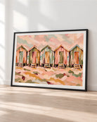 A framed art print depicting a row of colorful beach huts in a watercolor style, leaning against a white wall on a light wood floor.