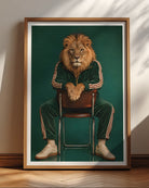 A framed art print featuring a lion with a human face sitting on a chair, wearing a green tracksuit with white stripes, set against a dark green background. The print is housed in a light brown wooden frame and leans against a beige wall on a wooden floor.