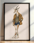 A framed art print featuring a whimsical fashion illustration of a gazelle dressed in casual clothing, leaning against a wall on a light wood floor.
