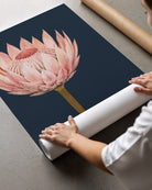 A person is unrolling a large wall art print or poster featuring a pink protea flower on a dark navy background, placed on a gray concrete floor with cardboard shipping rolls nearby.