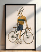 A framed art print leaning against a wall on a wooden floor. The print features a whimsical illustration of a gazelle with antlers dressed as a cyclist, wearing a yellow and green striped shirt, navy shorts, and holding a bicycle.