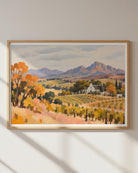 A framed landscape art print depicting a vineyard with autumn-colored trees, a small white church, and distant mountains, hung on a light gray wall with soft window shadows.