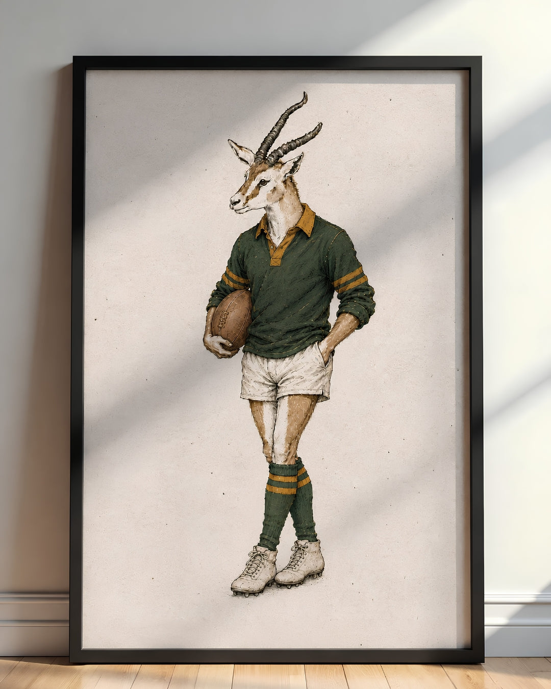 A framed art print featuring a whimsical illustration of a springbok antelope dressed in a vintage green and gold rugby jersey, shorts, and socks, leaning against a light gray wall on a wooden floor.