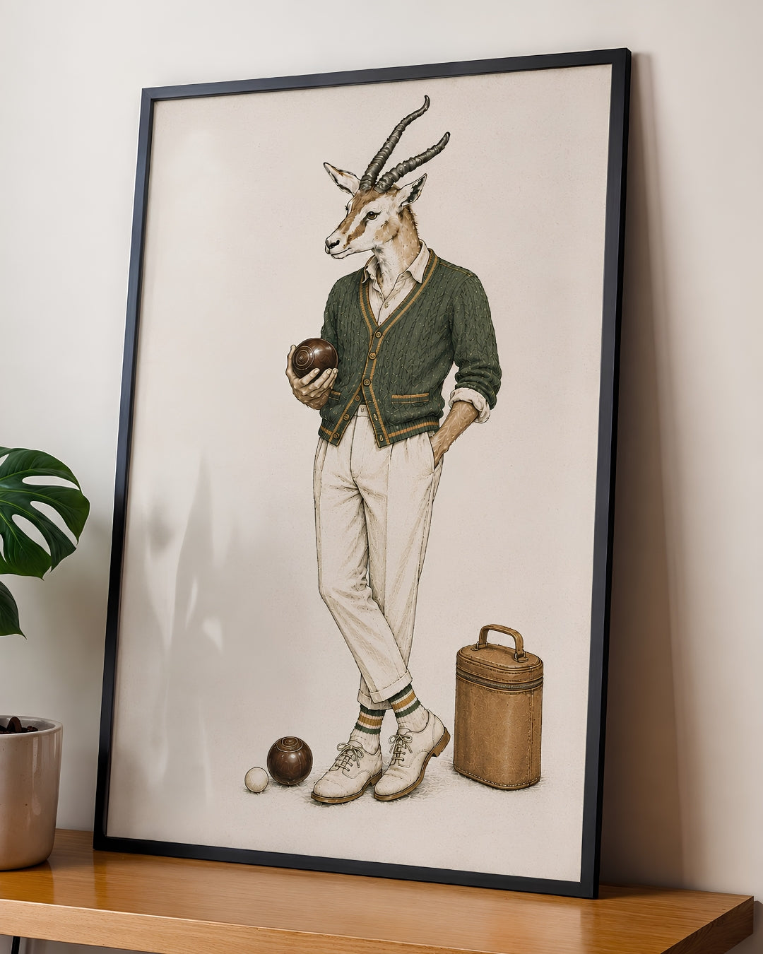A framed art print featuring a whimsical illustration of a gazelle dressed in vintage sportswear, leaning against a wall on a wooden shelf next to a potted plant.
