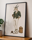 A framed art print featuring a whimsical illustration of a gazelle dressed in vintage sportswear, leaning against a wall on a wooden shelf next to a potted plant.