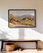 A framed landscape art print depicting an autumn vineyard with mountains in the background, displayed on a warm beige wall above a dark wood bench with a woven basket and shoes underneath.