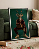A framed vertical art print leaning against a green sofa in a living room. The print shows a deer-like figure with antlers seated on a chair, wearing a red tracksuit against a dark teal background.