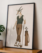 A framed art print featuring a whimsical illustration of a male antelope dressed in golf attire, leaning against a wall on a wooden shelf next to a potted plant.