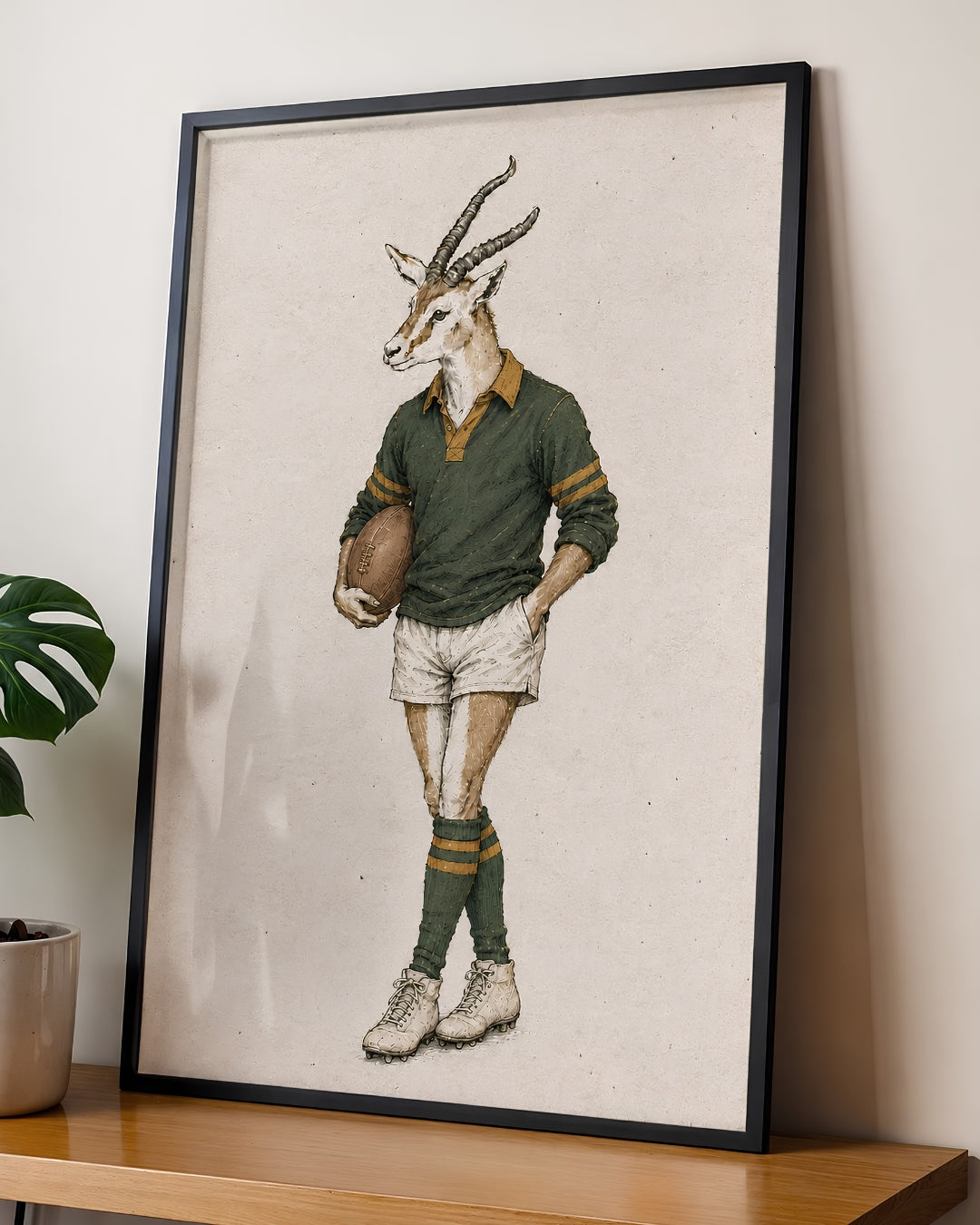 A framed art print featuring a vintage-style illustration of a springbok antelope dressed in a green rugby jersey, white shorts, and green socks, leaning against a wall on a wooden shelf next to a potted plant.