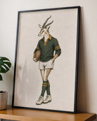 A framed art print featuring a vintage-style illustration of a springbok antelope dressed in a green rugby jersey, white shorts, and green socks, leaning against a wall on a wooden shelf next to a potted plant.