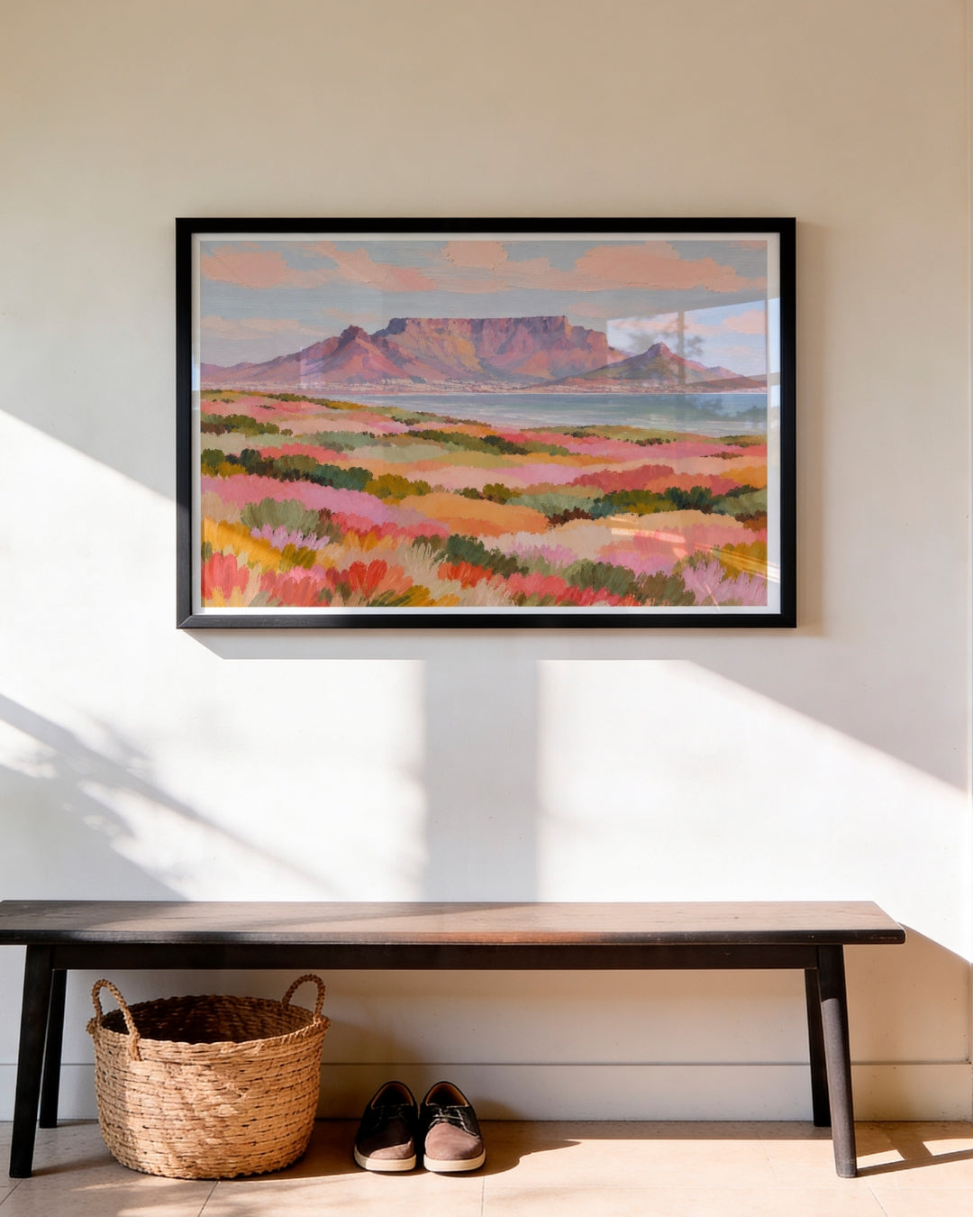 A framed landscape art print of a mountain and pink flower fields hangs on a beige wall above a dark wooden bench with a woven basket and shoes underneath.