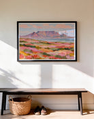 A framed landscape art print of a mountain and pink flower fields hangs on a beige wall above a dark wooden bench with a woven basket and shoes underneath.
