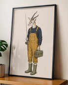 A framed art print featuring an anthropomorphic gazelle dressed in yellow overalls, a navy sweater, and green boots while holding a fishing rod and carrying a box, displayed on a wooden shelf against a beige wall.