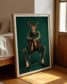A framed art print leaning against a bed in a warm, sunlit bedroom. The print features a whimsical illustration of a gazelle with antlers sitting on a chair, dressed in a green tracksuit against a dark teal background.