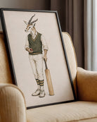 A framed art print featuring a whimsical illustration of a springbok antelope dressed in vintage cricket attire, leaning against a beige armchair in a styled interior setting.