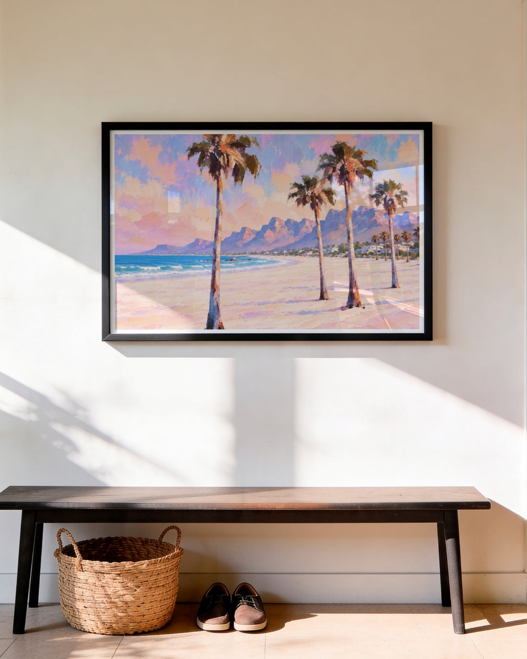 A framed landscape painting of a tropical beach with palm trees and mountains, displayed on a beige wall above a dark wooden bench with a woven basket and shoes underneath.