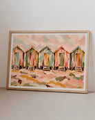 A framed watercolor painting of five beach huts in soft pastel colors, displayed against a neutral light gray wall and surface.