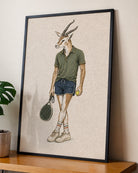 A framed art print featuring a whimsical illustration of a gazelle with a tennis head, wearing an olive green polo shirt, denim shorts, and holding a tennis racket and a tennis ball, displayed on a wooden shelf next to a potted plant.