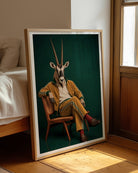A framed art print leaning against a wall in a sunlit bedroom. The print shows a stylized portrait of a gazelle with long horns seated in a chair against a dark green background, wearing a yellow tracksuit.