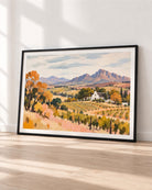 A framed landscape art print leaning against a white wall on a light wood floor. The artwork depicts a vineyard with rows of grapevines, orange and yellow trees, a small white farmhouse, and distant mountains under a cloudy sky.