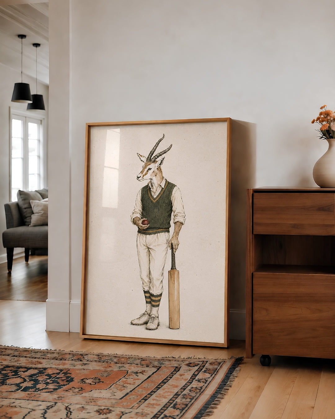 A framed art print featuring a whimsical illustration of a gazelle dressed in vintage cricket attire, leaning against a white wall in a modern living room setting.
