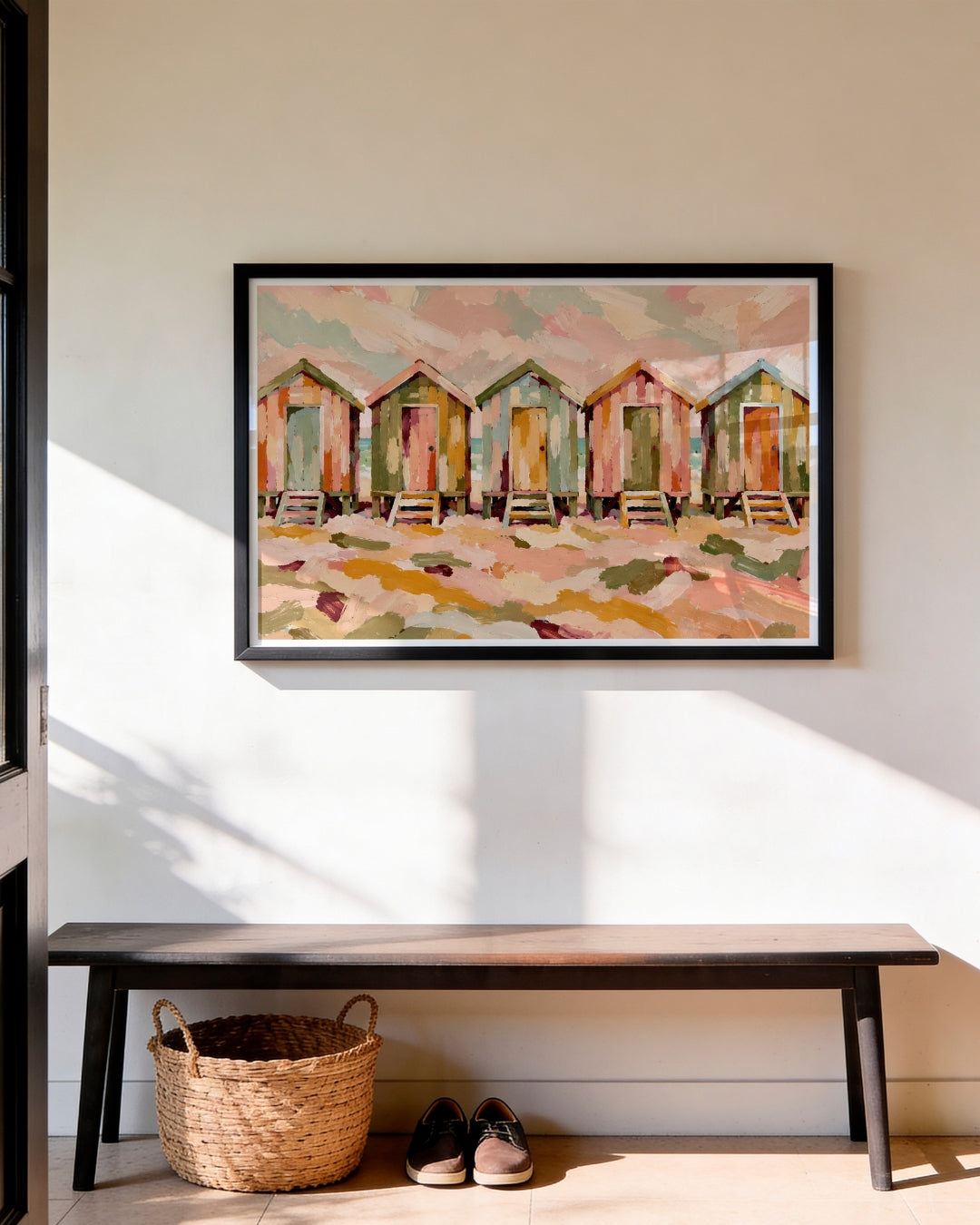 A framed painting of colorful beach huts hangs on a white wall above a wooden bench with a woven basket and shoes underneath.