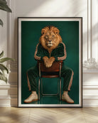 A framed art print featuring a lion dressed in a green tracksuit sitting on a chair, displayed leaning against a white paneled wall on a light wood floor next to a green plant.
