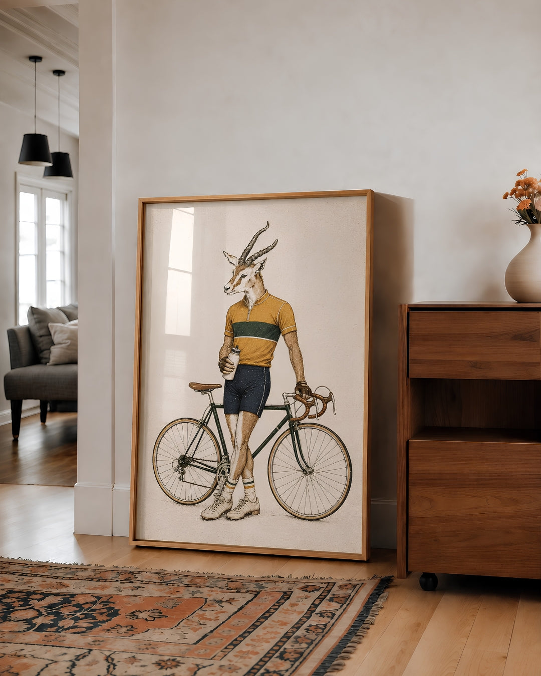 A large framed art print leaning against a white wall in a modern living room. The print features a whimsical illustration of a gazelle with antlers riding a vintage bicycle, wearing a yellow and green cycling jersey and navy shorts.