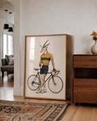 A large framed art print leaning against a white wall in a modern living room. The print features a whimsical illustration of a gazelle with antlers riding a vintage bicycle, wearing a yellow and green cycling jersey and navy shorts.