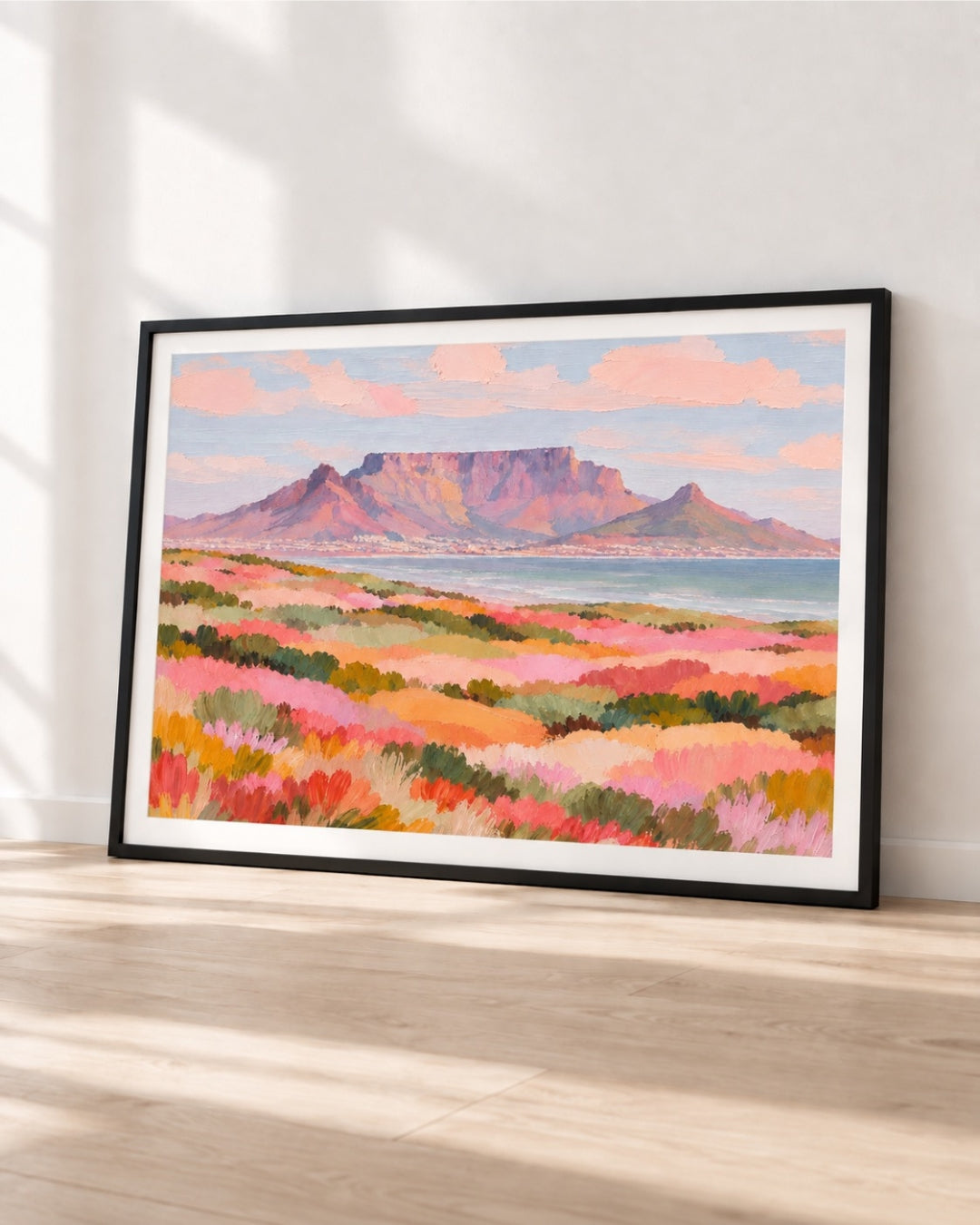 A framed landscape art print leaning against a white wall on a light wood floor. The artwork depicts a colorful coastal scene with pink and orange grasses in the foreground, a blue sea, and a mountain under a pink sky.
