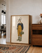 A framed art print featuring a whimsical illustration of a goat dressed as a farmer, standing in a rustic room next to a wooden cabinet.