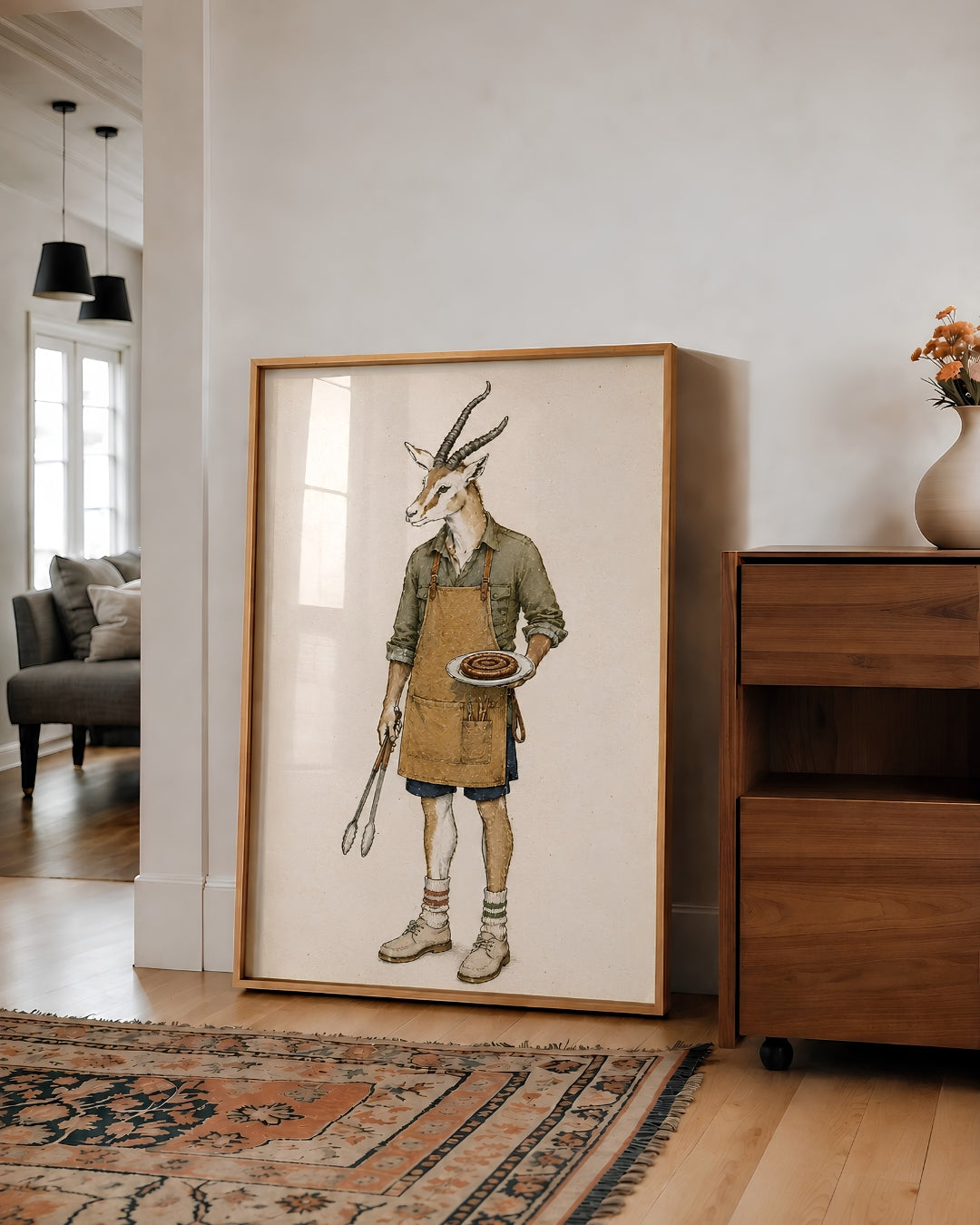 A large framed art print featuring a whimsical illustration of a gazelle dressed as a chef, leaning against a white wall in a modern living room next to a wooden cabinet.